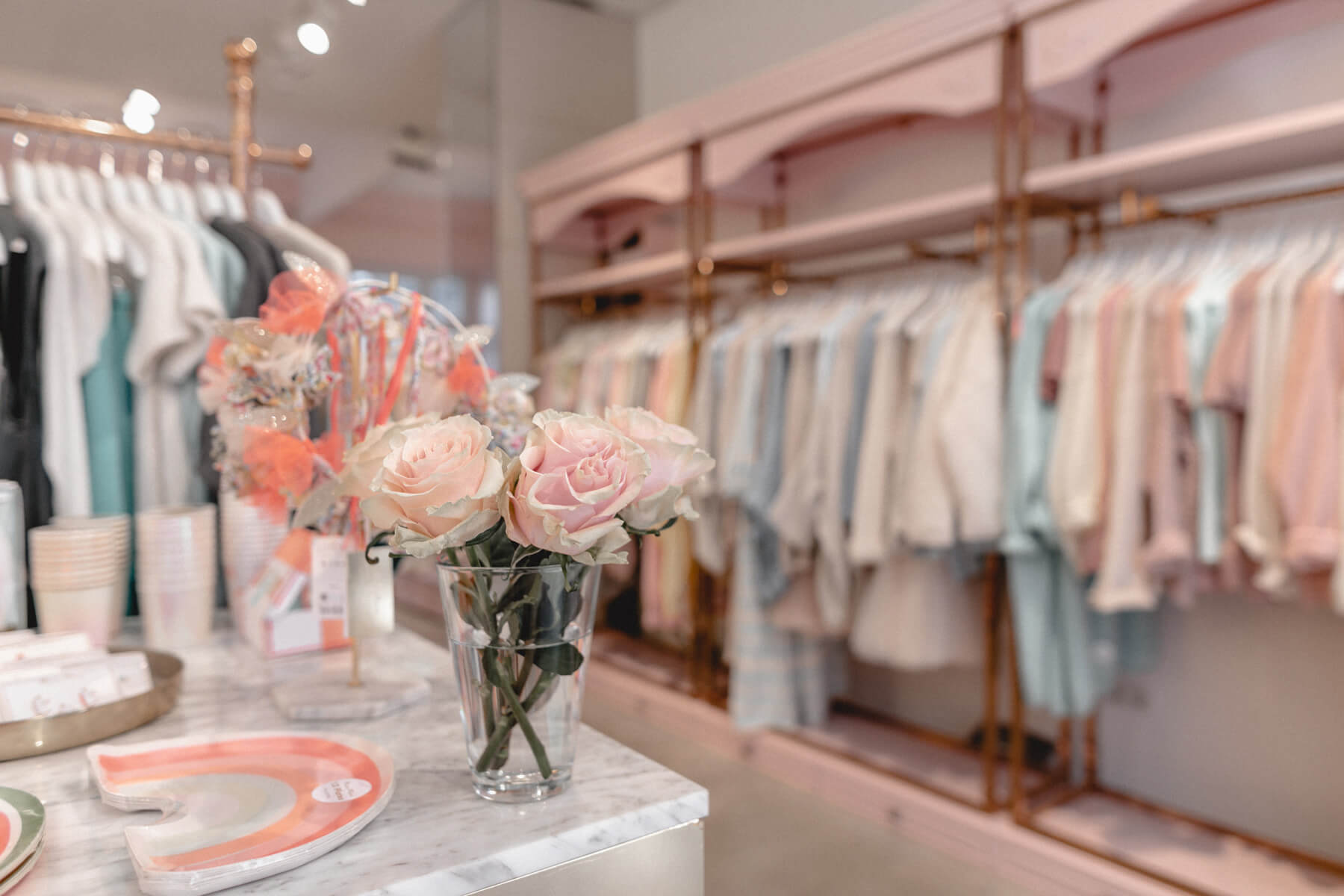 Blick in die Boutique Belgique: Kleiderstangen mit Damenmode, Geschenkartikel und ein Rosenstrauß auf der Marmortheke